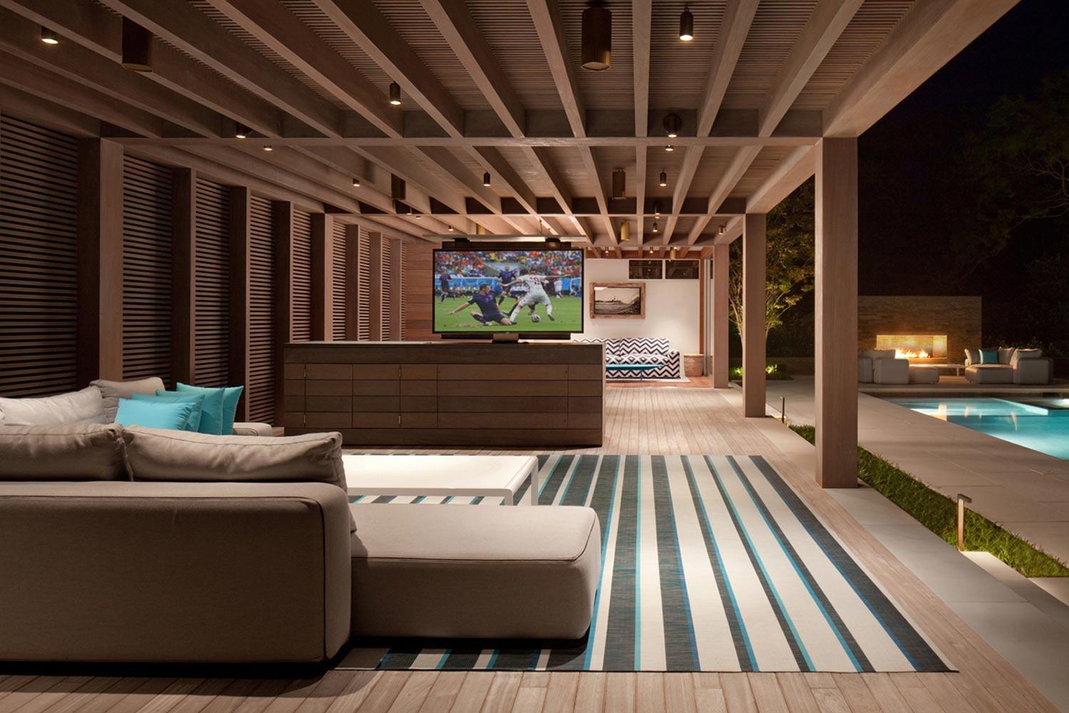 Luxurious outdoor lounge with a large TV, modern seating, and a poolside fireplace at night.