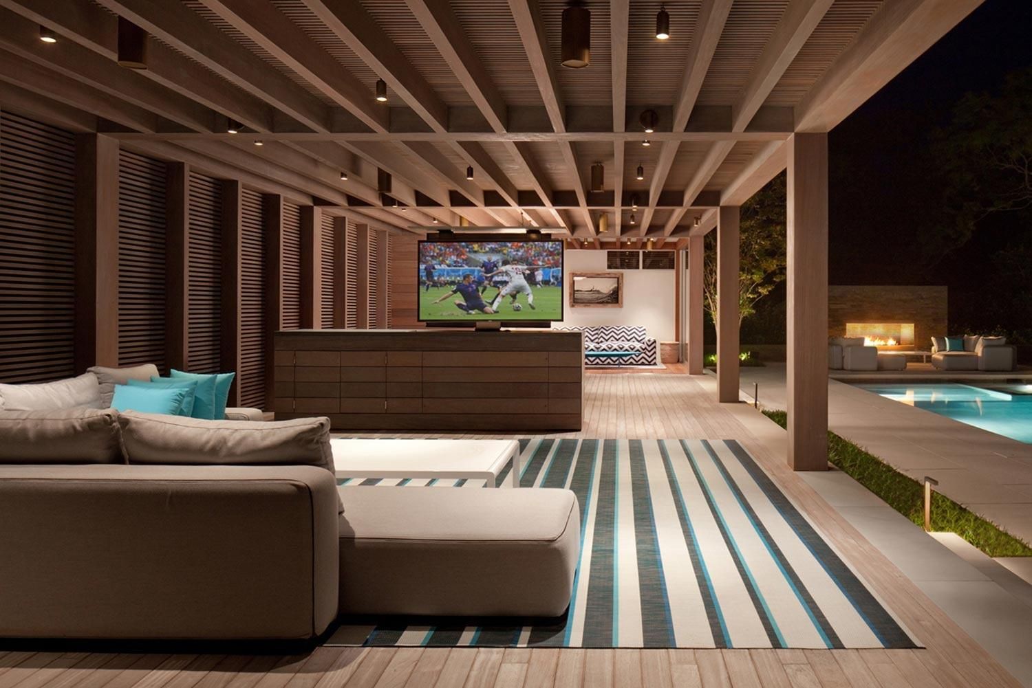 Luxurious outdoor lounge with a large TV, modern seating, and a poolside fireplace at night.
