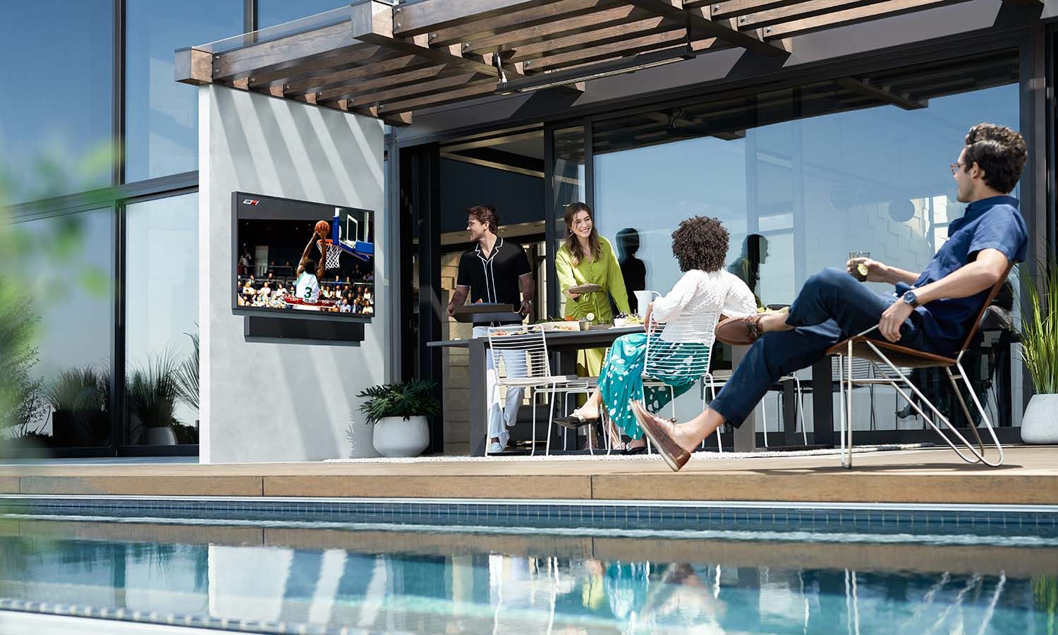 A group of people gathered in a backyard, enjoying a social gathering with outdoor seating, and a TV displaying a basketball game.